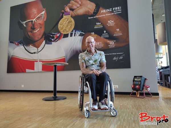 Heinz Frei im Rollstuhl vor einer Fotowand, auf der er selbst zu sehen ist und eine Goldmedaille hochhält
